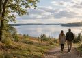 A couple walking along a wooded path towards a calm lake beneath soft September clouds in Denmark.