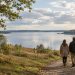 A couple walking along a wooded path towards a calm lake beneath soft September clouds in Denmark.